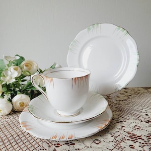 Pode incluir: Um conjunto de chá branco com xícara e pires com um padrão listrado verde e laranja. O conjunto está sobre uma toalha de renda branca.