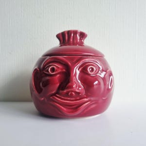 Könnte beinhalten: Ein glänzendes, tiefrotes Keramikgefäß mit Deckel, das auf der Vorderseite ein skulptiertes menschliches Gesicht zeigt. Das Gesicht hat markante Augen, eine Nase und einen lächelnden Mund. Der Deckel ist mit einem kleinen, gerüschten, runden Griff versehen, was dem Objekt eine bauchige Form verleiht.