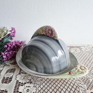 Könnte beinhalten: Graue Keramik-Butterdose mit einem Deckel in Helmform. Die Dose hat florale Akzente in Rosa, Gelb und Grün. Der ovale Boden und der geschwungene Griff sind sichtbar. Die Dose steht auf einer weißen Spitzendecke. Maße in cm.