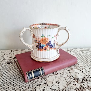Pode incluir: Uma caneca de cerâmica branca com duas alças e um desenho floral em laranja, azul e castanho. A caneca tem uma textura canelada e acabamento dourado. Está em cima de um livro bordô com letras douradas.