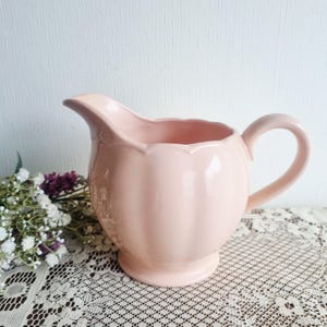 May include: A pale pink ceramic creamer jug with a scalloped edge and a curved handle. The jug has a round base and a spout. It is displayed on a lace tablecloth, with flowers in the background.