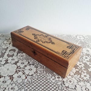 Op de afbeelding: Een rechthoekige houten kist met een scharnierend deksel. De kist is lichtbruin met een donkerbruin geometrisch ontwerp rond de randen en het woord "GLOVES" in het midden. De kist staat op een wit kanten tafelkleed.