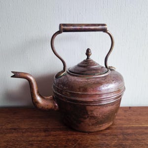 Peut inclure: Ancienne bouilloire en cuivre avec un bec courbé et une poignée. La bouilloire a un couvercle avec un petit bouton et est posée sur une surface en bois. Le cuivre est de couleur brun-rouge et montre des signes d'âge.