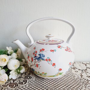 Pode incluir: Bule de porcelana branca com um design floral com flores vermelhas e pássaros azuis. A chaleira tem uma borda dourada em torno da tampa e uma banda dourada em torno do corpo.