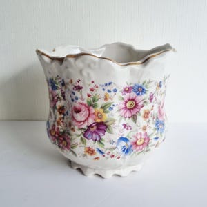 May include: A white ceramic planter with a scalloped rim and base, featuring a colourful floral pattern of pink, purple, yellow, and blue flowers with green leaves. The top edge of the planter is accented with a thin, metallic gold painted trim.