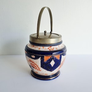 May include: A vintage ceramic biscuit jar with a silver-toned metal lid and handle. The jar has a cream base with blue and orange floral and geometric designs. The handle is arched, and the lid has a small knob.