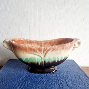 Pode incluir: Um pequeno vaso de cerâmica oval com duas alças. O vaso é esmaltado em tons de marrom, verde e branco, com um efeito manchado. Tem uma base marrom.