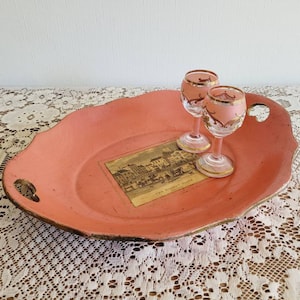 May include: A pink oval serving tray with gold trim and two handles. The tray features a small vintage postcard that reads "The Market Place". Two clear glass shot glasses with gold trim are on the tray.
