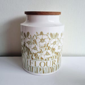 以下が含まれることがあります： 木製の蓋が付いた白いセラミック製の小麦粉容器。容器には金色の花の模様と、金色の文字で「FLOUR」の文字が描かれています。デザインは容器を囲んでいます。容器は円筒形で、底が少し広くなっています。
