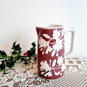 Peut inclure: Un pichet en céramique rouge avec un motif floral en relief blanc. Le pichet a une anse et une large ouverture.