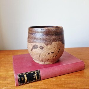 Könnte beinhalten: Ein handgefertigter Keramikbecher mit rustikalem, gesprenkeltem Aussehen. Die obere Hälfte ist dunkelbraun, geht in ein helleres Beige mit braunen Sprenkeln über. Der Becher steht auf einem geschlossenen, roten Buch mit goldener Schrift.