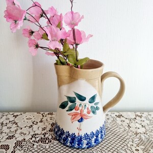 Op de afbeelding: Een keramische kan met een bruin top en handvat, en een witte body versierd met een bloemenontwerp in blauw, roze en groen. Roze bloemen zijn in de kan gerangschikt. De kan staat op een wit kanten tafelkleed.