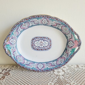 May include: An oval serving platter with a white background and a colorful floral design. The platter has two handles and a gold rim.