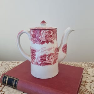Peut inclure: Une cafetière en céramique blanche et rouge avec un couvercle, une poignée et un bec. La cafetière présente un motif floral et paysager rouge. La cafetière est posée sur un livre rouge.