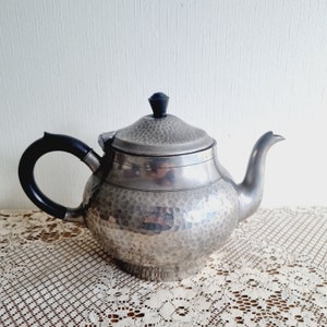 May include: A silver hammered teapot with a black handle and knob on the lid. The teapot is sitting on a white lace tablecloth.