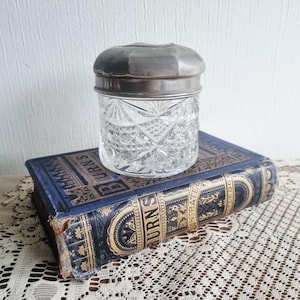 Könnte beinhalten: Ein klares Glas mit einem silberfarbenen Deckel. Das Glas hat ein geometrisches Muster und steht auf einem Vintage-Buch mit blauem Einband und goldener Schrift. Das Buch liegt auf einer cremefarbenen, spitzenartigen Oberfläche.