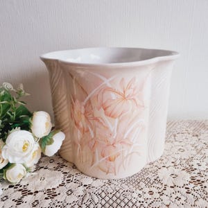 May include: A white ceramic planter with a scalloped rim and a floral design featuring pink and orange flowers. The planter has a textured surface and a wavy design.