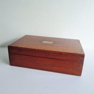 May include: A rectangular wooden box with a reddish-brown finish and a visible wood grain. The lid features a centered, rectangular brass inlay plate. The box is constructed with small metal screws visible along the side and front edges.