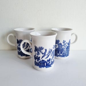 May include: Three white ceramic mugs with blue Willow pattern illustrations. The mugs have a cylindrical shape with a curved handle. The blue design depicts a landscape with buildings, birds, and a boat.