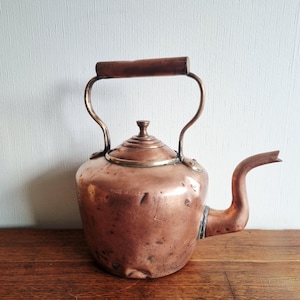 Puede incluir: Una tetera de cobre antigua con asa de madera y boquilla curva. La tetera tiene un cuerpo redondeado y una tapa con un pequeño pomo. El cobre tiene un color marrón rojizo cálido, y el asa es de color marrón oscuro.