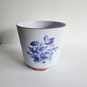 May include: A white ceramic planter featuring a blue floral pattern on the front. The vessel has a tapered shape with a gold-coloured rim and a gold-coloured base. The floral design consists of various blossoms and leaves in shades of blue.