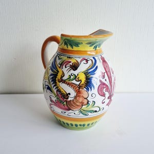 Könnte beinhalten: Keramikkrug mit handbemaltem Design. Der Krug hat einen gebogenen Griff und Ausguss mit einem farbenfrohen Muster aus einem Fabelwesen, floralen Elementen und dekorativen Bordüren in Gelb-, Blau-, Rot- und Grüntönen.