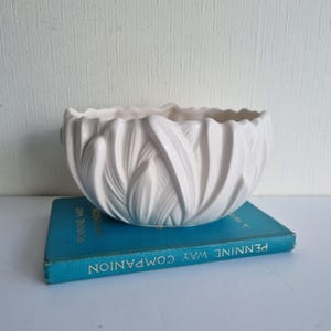 May include: A white ceramic bowl with a textured, leaf-like design. The bowl is placed on a vintage blue book titled "Pennine Way Companion". The bowl's rim has a scalloped edge, a decorative element for home decor.