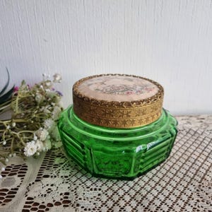 May include: A vintage green glass jar with a decorative lid. The lid is round with a gold-colored metal rim and a fabric top featuring a floral design. The jar has a faceted, octagonal shape.