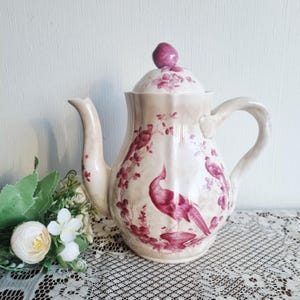 May include: Cream-colored ceramic teapot with a lid and handle, featuring a maroon peacock and floral design. The teapot has a curved spout and a rounded handle. The lid is topped with a maroon knob. The teapot is on a lace tablecloth.