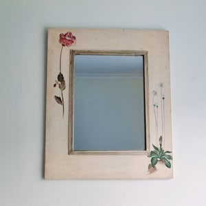 May include: A white wooden framed mirror with a painted floral design. The design features a red poppy and white flowers with green leaves.