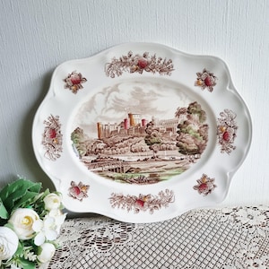 May include: A white, scalloped-edge serving platter with a detailed landscape scene of a castle in shades of brown, red, and green. Floral and foliage designs in brown and red accent the rim.