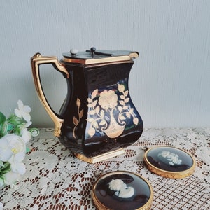 May include: A black and gold ceramic pitcher with a floral design and a lid. Two small round frames with gold trim and black backgrounds contain cameo portraits.