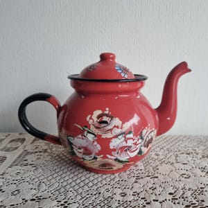 Pode incluir: Uma chaleira de esmalte vermelho com um design floral pintado à mão com pinceladas brancas e castanhas no corpo. A chaleira tem uma pega preta, uma tampa vermelha a condizer com pequenos detalhes florais e um rebordo preto.