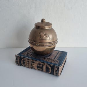 May include: A spherical brass container with a removable lid, featuring an embossed lion figure and the text "BRITISH EMPIRE EXHIBITION 1924" engraved around the centre. The container rests on a rectangular, dark blue and gold antique book.