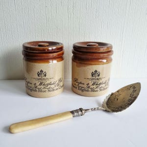 May include: Two antique ceramic jars with brown lids and labels, and a silver and ivory cheese server. The jars are labelled "Paxton & Whitfield Ltd. Finest English Blue Stilton Cheese."