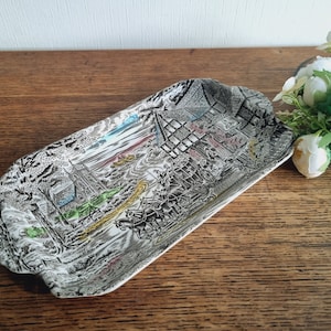 Może przedstawiać: Prostokątne ceramiczne naczynie do serwowania z czarno-białym wzorem przedstawiającym scenę wiejską z kościołem, domami i ludźmi. Naczynie ma podwyższony rant i lekko zakrzywiony kształt.