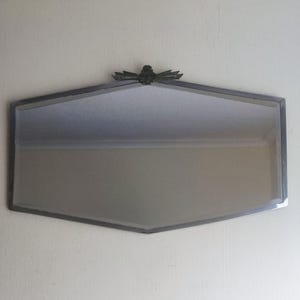 Pode incluir: Espelho vintage em estilo Art Déco, com formato octogonal. Possui moldura de metal prateado e um emblema decorativo no topo. Está montado em uma parede creme. Dimensões: aproximadamente 60 cm x 40 cm.