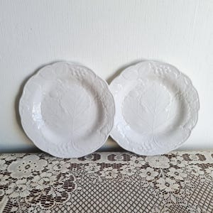 Può includere: Due piatti in ceramica bianca con un motivo a rilievo di foglie e uva. I piatti hanno un bordo smerlato e sono appoggiati su una tovaglia di pizzo bianco. I piatti sono di design classico.