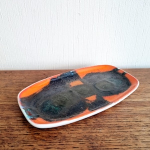 May include: A small, rectangular ceramic dish with a white rim and an orange glaze. The dish has a black and green abstract design.