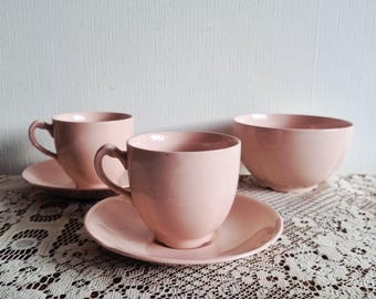 May include: A set of three pink ceramic dishes. Two teacups with saucers and a bowl are arranged on a lace tablecloth. The teacups have curved handles and the bowl has a rounded shape. The dishes are a solid, soft pink color.