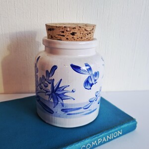May include: A white ceramic jar with a cork stopper, decorated with blue floral patterns. The jar has a cylindrical shape with a wider base and a narrow neck. The blue floral design features stylized leaves and flowers. The jar is placed on a blue book.