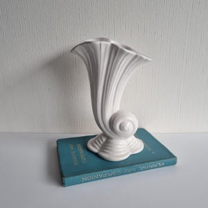 Puede incluir: Un jarrón de cerámica blanco con diseño de concha. El jarrón tiene una parte superior acampanada y una base curva en espiral. Se encuentra sobre un libro verde azulado. Es un objeto decorativo.