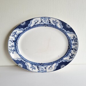 May include: Oval serving platter with a white center and a blue decorative border. The border features a repeating pattern of figures, trees, and architectural elements. The platter has a classic, vintage aesthetic.