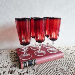 May include: Set of six vintage red-tinted stemmed glasses. The glasses have a clear stem and base, with a deep red color that fades to a darker shade at the rim. They are arranged on a red book with gold lettering.