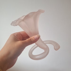 Könnte beinhalten: Eine Skulptur aus mattiertem, hellrosa Glas einer Calla-Lilie. Die trompetenförmige Blüte der Blume krümmt sich zu einem anmutigen, spiralförmigen Stiel. Die Skulptur wird vor einem schlichten, weißen Hintergrund gehalten.