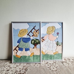 May include: Two framed fabric wall art pieces featuring quilted scenes of figures in straw hats. One figure carries a ladder and basket, while the other holds a flower. The designs use blue, yellow, green, and white fabric patches with stitched details.
