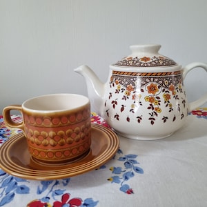 May include: A brown and orange patterned teacup and saucer set with a matching white teapot with a floral design. The teapot has a brown and orange floral pattern with a white background.