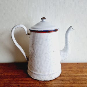 May include: White enamel teapot with a blue and red rim. The teapot has a textured surface and a rounded body.
