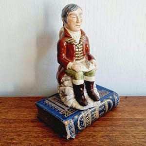 以下が含まれることがあります： 赤いコートと緑の半ズボンを着て、紙を持っている座っている男性の陶器製の置物、青い本の上にあります。置物は塗装された特徴で詳細に描かれており、石のような台座に座っています。本には金色の文字が書かれています。