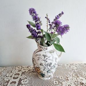 以下が含まれることがあります： 黒と白の植物柄に淡い黄色のアクセントが施された白い陶器製のピッチャー。紫色のラベンダーの枝のブーケが生けられています。ピッチャーには曲線的なハンドルと広い注ぎ口があり、詳細な花のイラストが描かれています。